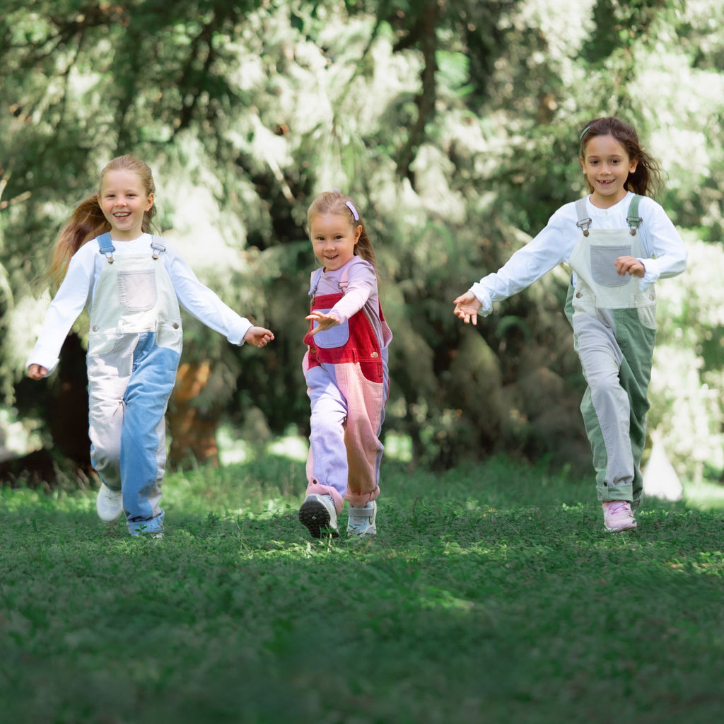 Three children running outdoors in a forested area wearing dungarees